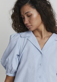 Camicia azzurro chiaro a maniche corte con tessuto texture, maniche a sbuffo e colletto a forma di V. Bottoni discreti completano il design.