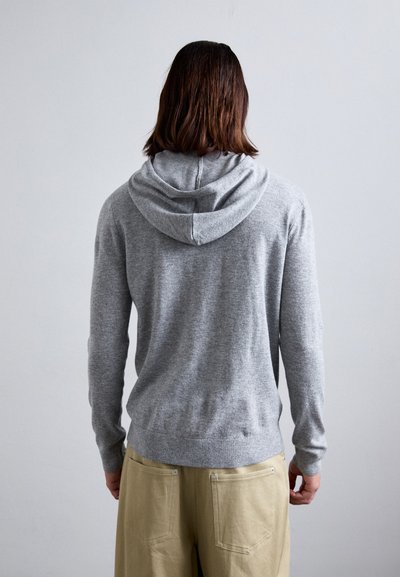 Hellgrauer Hoodie mit weicher Textur, körpernahem Schnitt und Kängurutasche, über beigen Hosen getragen, von hinten betrachtet.