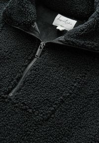 Pull en polaire noir avec surface texturée, fermeture éclair métallique demi-zip, et étiquette blanche à l'intérieur du col indiquant « downtime NEXT ».