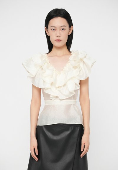 Femme portant un chemisier blanc transparent à volants avec un décolleté en V et une jupe noire, se tenant les bras relâchés contre un fond blanc uni.