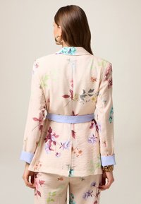 Blazer fleuri en tissu texturé de pêche clair avec des manchettes et une ceinture bleues ; présente divers motifs floraux colorés dans tout le vêtement.