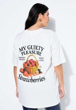 Kobieta o długich ciemnych włosach ubrana w biały, za duży T-shirt z koszykiem truskawek i napisem „My Guilty Pleasure Strawberries” na plecach.