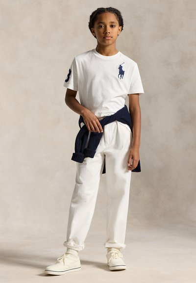 Copil purtând tricou polo alb cu logo bleumarin, pantaloni albi, adidași crem și pulover bleumarin legat în jurul taliei, stând pe un fundal neutru.