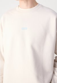 Persona che indossa un maglione beige chiaro a girocollo con un piccolo logo blu "HUGO" centrato sul petto.