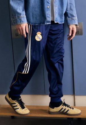 Persona con pantaloni sportivi del Real Madrid blu navy, scarpe da ginnastica Adidas beige e giacca di jeans in piedi su una panchina di legno.