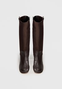 Une paire de bottes noires en cuir jusqu'aux genoux avec des parties en cuir sur le pied et des tiges en tissu, se tenant debout sur un fond clair.