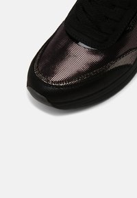 Pointe de chaussure de sport noire avec un revêtement en maille foncée brillante et une zone de lacets noire unie sur un fond blanc.