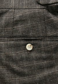 Grau karierter Stoff mit strukturiertem Oberflächen, neben einer Nahtdetails befindet sich ein cremefarbener Knopf mit vier Löchern.
