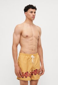 Gelbe Badehose mit einem roten Wellendesign am Saum, ausgestattet mit einem weißen Kordelzug, aus leichtem, schnell trocknendem Material.