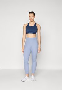 Jūrų pajėgų sportinė liemenėlė su logotipo juostele, derinama su šviesiai mėlynomis aukštu liemeniu legging'ais. Modelis dėvi baltus sportinius batus. Lygus paviršius visame gaminyje.