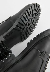 Zapatos de cuero negro con una suela de goma texturizada que presenta tacos prominentes y patrones geométricos. Costura reforzada a lo largo de la parte superior.