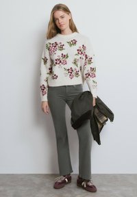 Suéter tejido con patrón floral en burdeos y verde, combinado con pantalones acampanados grises y zapatillas oscuras. La modelo sostiene una chaqueta.
