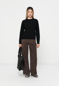 Svart långärmad tröja, bruna manchesterbyxor med vida ben, håller en svart tote-väska och bär grå sneakers mot en enfärgad bakgrund.