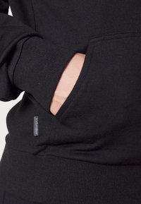 Sudadera negra con bolsillo canguro. El material es suave y texturizado, con puños acanalados y una pequeña etiqueta en la costura del bolsillo.