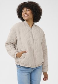 Veste bomber matelassée beige avec fermeture éclair devant, poignets côtelés, poches latérales et ourlet élastique. Texture lisse avec un motif en diamant.