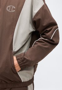 Άνδρας που φοράει καφέ και μπεζ μπουφάν με λευκό περίγραμμα, το χέρι στην τσέπη του μπουφάν, με εμφανές το λογότυπο της Champion στο στήθος.