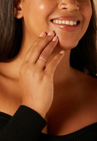 Gold ring with three clear gemstones, set on a model's finger while she lightly touches her cheek. Soft lighting enhances details.