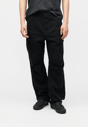 Persona que lleva pantalones cargo negros holgados y zapatos negros sin cordones, de pie contra un fondo claro y sencillo.