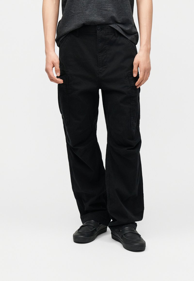 Persona que lleva pantalones cargo negros holgados y zapatos negros sin cordones, de pie contra un fondo claro y sencillo.