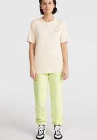 Camiseta de color beige claro con mangas cortas y un logotipo texturizado en el pecho, combinada con joggers de color verde lima brillante y zapatillas negras.
