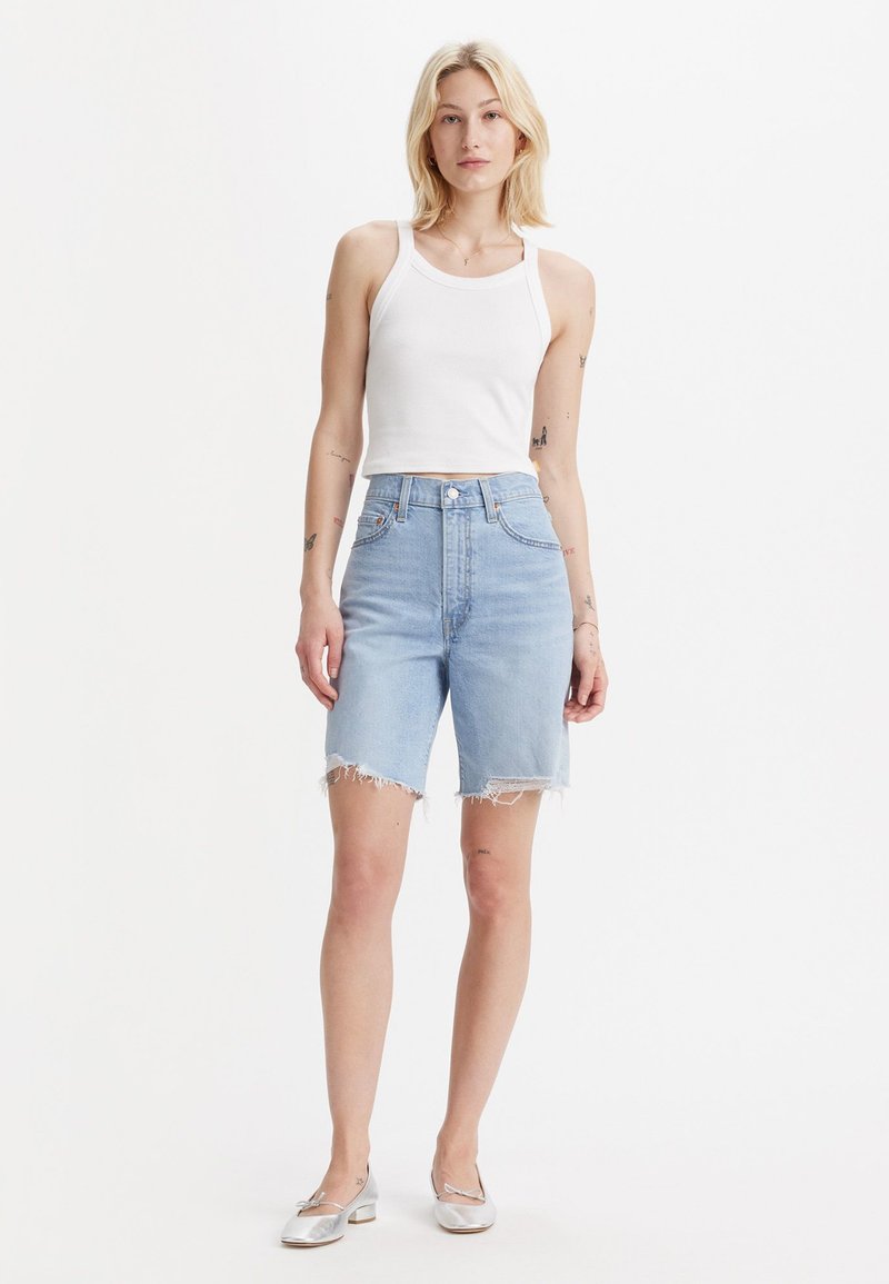 Shorts en denim bleu clair avec des ourlets effilochés, taille haute, associés à un débardeur côtelé blanc et des ballerines argentées. Design minimaliste.