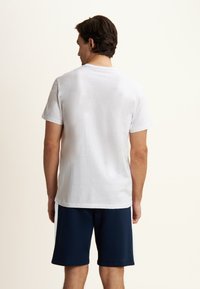 T-shirt bianco in cotone con maniche corte, design semplice, indossato con pantaloni corti blu navy, vista da dietro. Tessuto leggero, senza motivi visibili.
