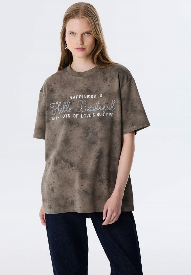 Olijfgroene oversized t-shirt van zachte stof, met een verouderde textuur en witte geborduurde tekst met glinsterende accenten.