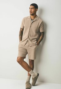 Beige short-sleeve button-up shirt with ribbed texture and matching shorts. Effortless fit, accented by brown slip-on shoes and beige socks.