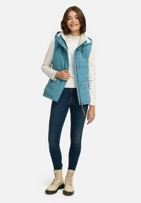 Light blue padded vest with hood, zip and snap closures, worn over a white sweater and dark blue jeans, paired with cream lace-up boots.