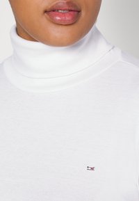 Pull à col roulé blanc en tissu côtelé, doté d'un petit logo sur la poitrine gauche. Texture douce avec une coupe ajustée.