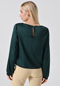 Donkergroene satijnen blouse met lange mouwen, keyhole detail aan de achterkant vastgemaakt met een knoop, zachte textuur en een licht cropped pasvorm.