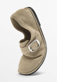 Beige suede shoe with soft texture, rounded toe, single buckle strap, and black rubber sole. Features minimalistic design and understated style.