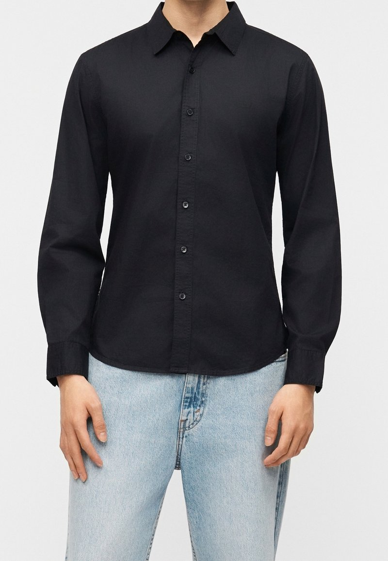 Personne portant une chemise noire unie à manches longues boutonnée avec un col et un jean bleu clair, debout devant un fond blanc.
