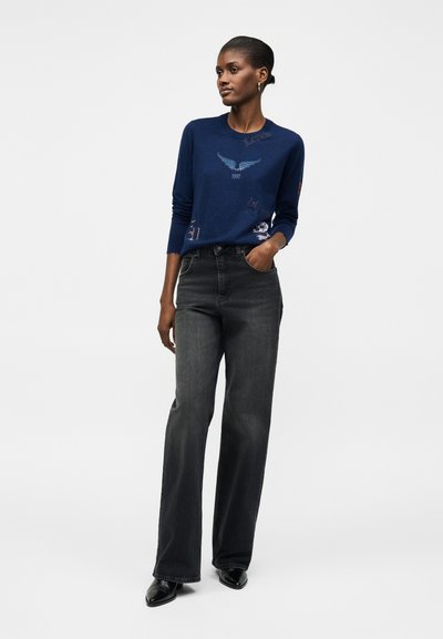 Γυναίκα που φοράει μπλε σκούνο πουλόβερ με σύμβολα και μαύρο παντελόνι καμπάνα με ψηλή μέση, στέκεται με το ένα χέρι στην τσέπη σε λευκό φόντο.