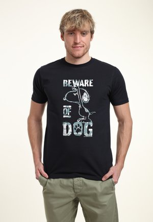 Ung mand iført en sort t-shirt med en tegneseriehund og teksten "Beware of dog,", kombineret med lysegrønne bukser, står med hænderne i lommerne.