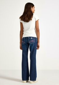 Mørkeblå vidde jeans med lyserød syning på baglommerne og en lysefarvet t-shirt, set bagfra.