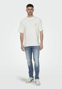 Romige katoenen oversized t-shirt met korte mouwen en een subtiele bedrukte logo op de linkerborst, gecombineerd met lichtblauwe versleten jeans en witte sneakers.