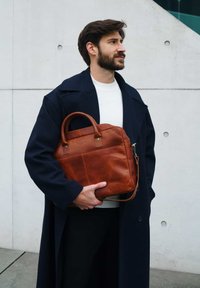 Brown leather bag with top handles, smooth texture, and a detachable strap; features a front pocket and subtle logo detail; worn with a navy coat.