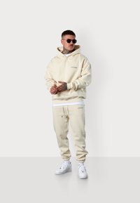 Cremefarbener Hoodie und Jogginghose mit elastischen Bündchen, mit einem Logo auf der Brust und an der Seite. Getragen mit weißen Sneakern und Sonnenbrille.