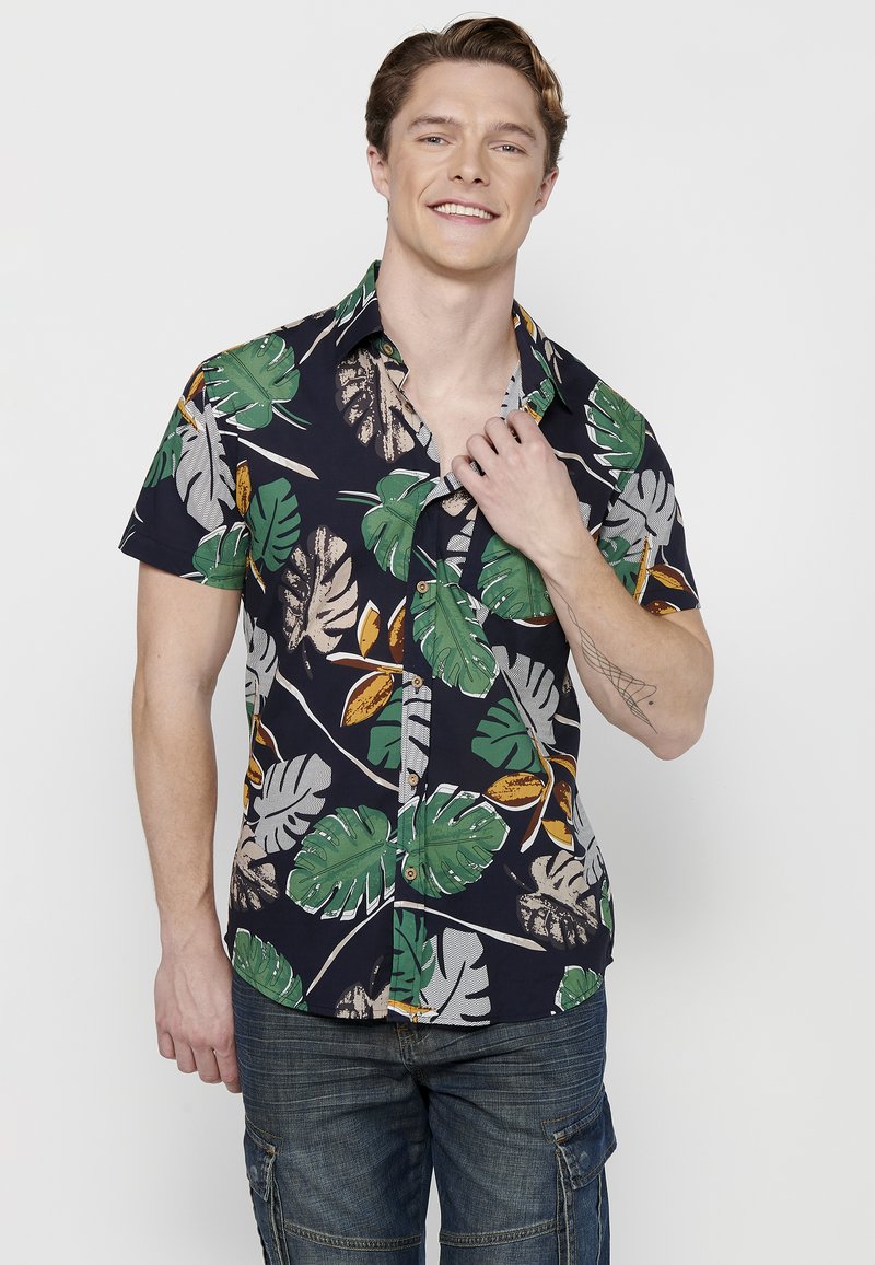 Camisa de manga corta con fondo azul marino oscuro y patrones de hojas verdes y beige. Estilo de botonadura con cuello puntiagudo.