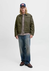 Mand iført olivengrøn jakke, rutet skjorte, bleget blå jeans, brune sko og marineblå kasket, står op ad en ensfarvet baggrund.