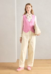 Vesta tricotată roz cu buzunare, bluză albă, pantaloni largi bej și pantofi roz cu o talpă texturată, ținând o geantă bej.