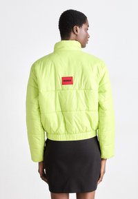 Veste matelassée vert fluo à design court et bord élastiqué. Présente un logo "HUGO" rouge bien visible au dos. Texture lisse et rembourrée.