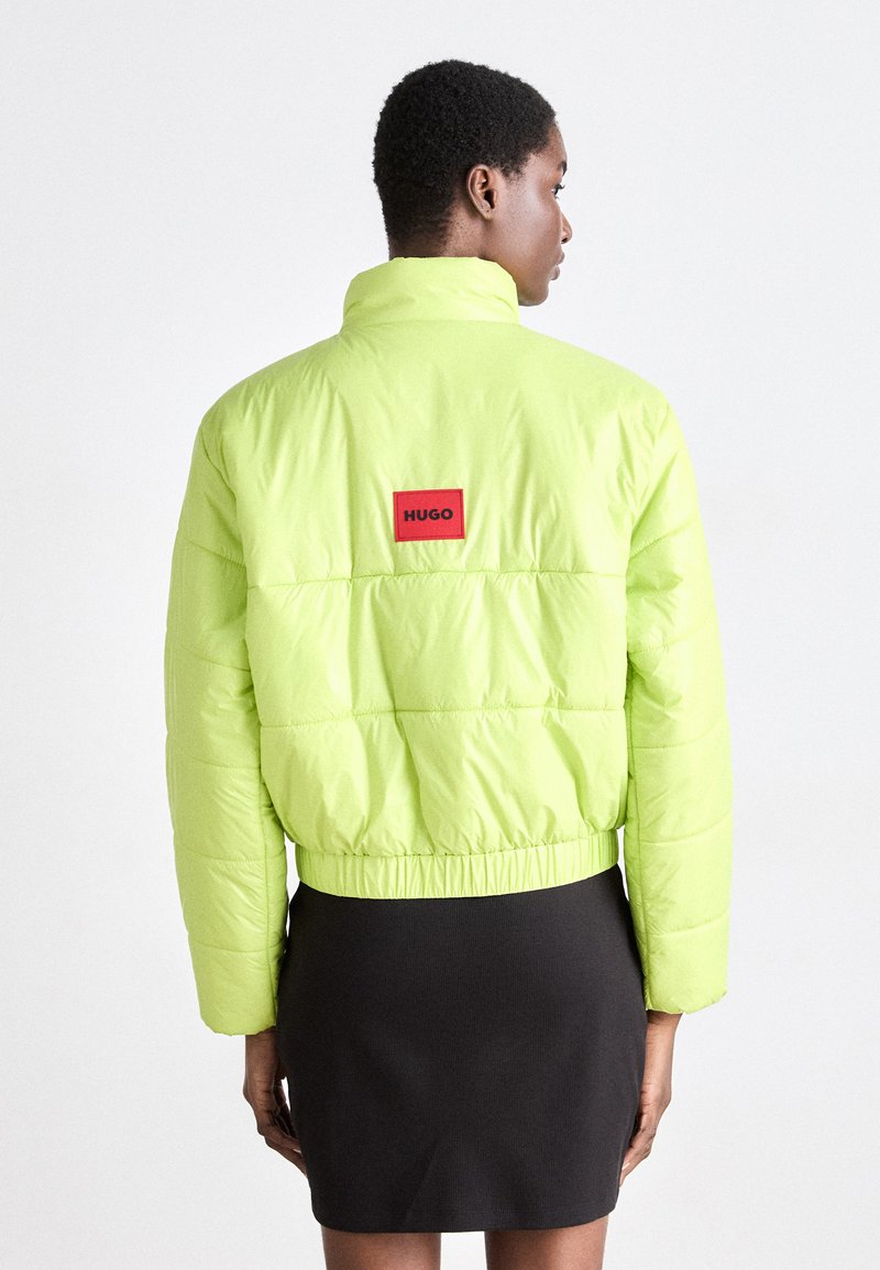 Veste matelassée vert fluo à design court et bord élastiqué. Présente un logo "HUGO" rouge bien visible au dos. Texture lisse et rembourrée.