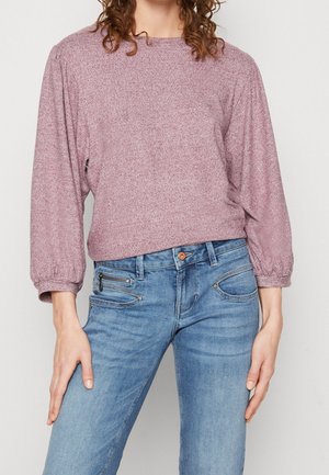 Femme portant un pull à manches longues violet clair et un jean en denim bleu avec des détails de fermeture éclair et boutons, debout devant un fond uni.
