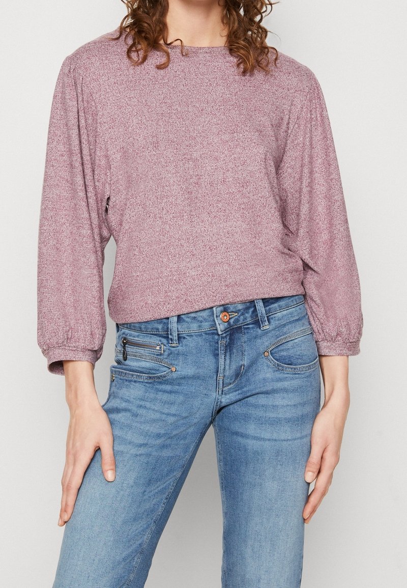 Femme portant un pull à manches longues violet clair et un jean en denim bleu avec des détails de fermeture éclair et boutons, debout devant un fond uni.
