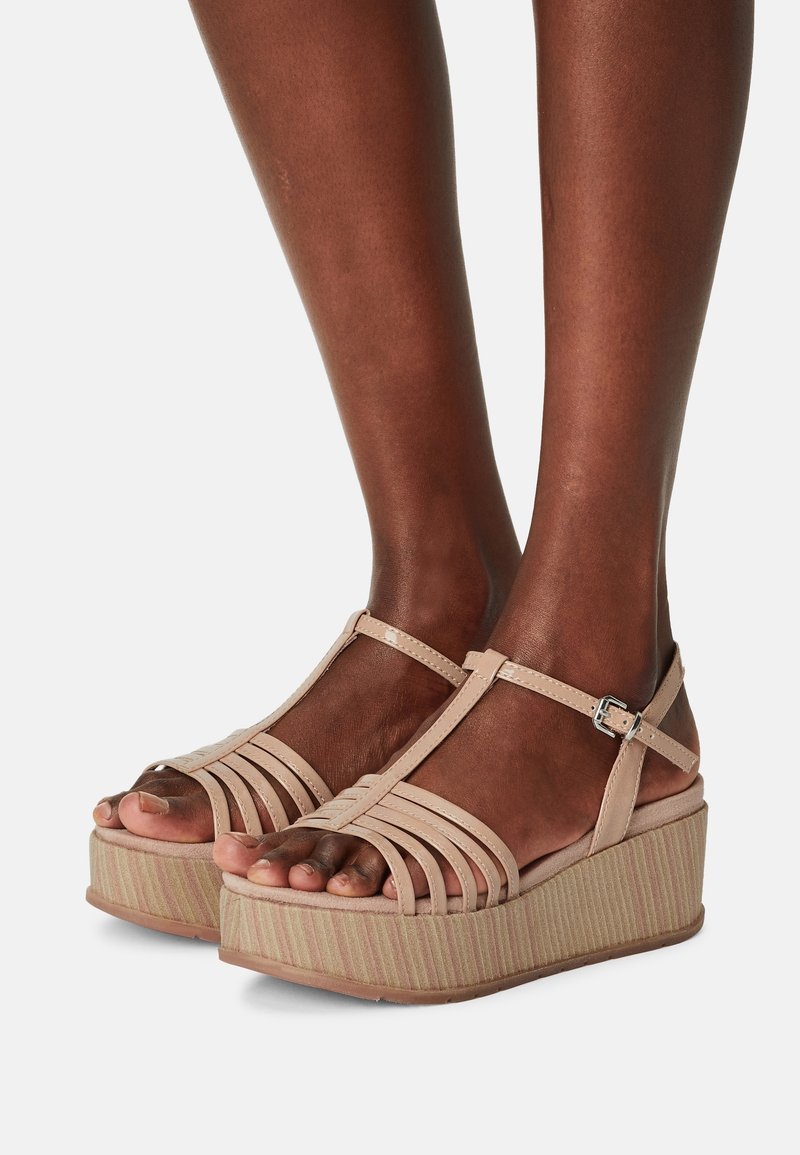 Jambes à la peau foncée portant des sandales compensées beiges à lanières avec boucles réglables à la cheville sur un fond clair et neutre.