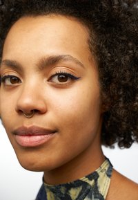 Primo piano di una persona con capelli ricci, che indossa un top senza maniche a fantasia. Le caratteristiche includono occhi marroni con eyeliner blu e un anello al setto oro.