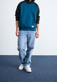 Felpa teal e nera con tasca frontale, abbinata a jeans baggy azzurri e sneakers bianche su una superficie in moquette blu.