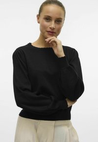 Czarny dzianinowy sweter z falbaniastym dekoltem i bufiastymi rękawami. Miękka tekstura oraz dopasowany krój, w zestawie z jasnymi spodniami.